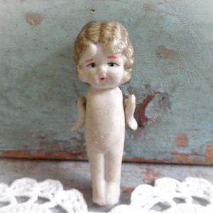 May include: A small, vintage bisque doll with a painted face and curly blonde hair. The doll has a sad expression with red accents on the lips and eyebrows. The doll is nude and has small arms and legs. The background is a distressed blue surface.