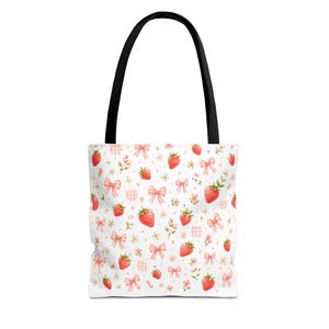 Puede incluir: Un bolso tote blanco con asas negras que presenta un patrón repetitivo de fresas rojas, pequeños lazos rosas, diminutas flores blancas y cuadrados a cuadros de color rosa claro dispersos por la superficie en un estilo de ilustración de acuarela.