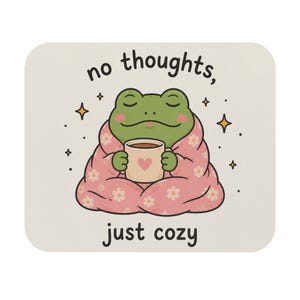 Puede incluir: Una alfombrilla de ratón rectangular con fondo beige claro que muestra una rana verde envuelta en una manta rosa con flores, sosteniendo una taza beige con un corazón rosa. El texto superior dice no thoughts, y el inferior just cozy.