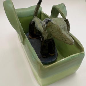 May include: A ceramic dog sculpture in a green rectangular container. The dog is black and green with a long body and ears. The container has a light green exterior and a darker green interior.