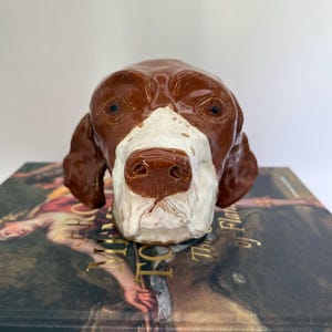 May include: A ceramic dog head sculpture with brown and white markings. The sculpture features a detailed nose, eyes, and ears. The dog head is resting on a book with gold lettering and a painting on the cover.