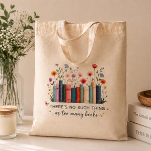 May include: A beige canvas tote bag featuring a colourful graphic of a row of books with wildflowers growing from the top. The text "THERE'S NO SUCH THING as too many books" is printed in black below the books, accompanied by small butterflies.