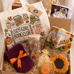 May include: A gift box containing a cream-coloured tote bag with "COZY GIRL BOOK CLUB" text, a glass tumbler with a bamboo lid and autumn-themed decals, a dark purple folded cloth with an orange ribbon, a pumpkin candle, and a crocheted sunflower.