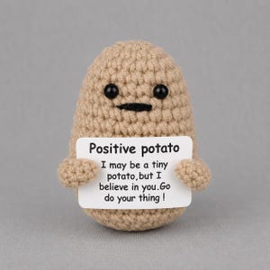 May include: A tan crocheted potato figure with two black bead eyes and a black stitched mouth. It holds a white rectangular sign that reads: Positive potato. I may be a tiny potato, but I believe in you. Go do your thing!