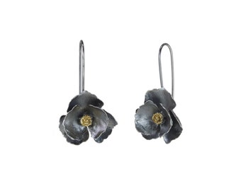Puede incluir: Un par de pendientes colgantes con pétalos de flor en tono plata oxidada oscura y un centro metálico texturizado en tono dorado. Cada pendiente cuelga de un gancho de alambre largo y fino en tono plata, creando un diseño floral con un contraste de acabados metálicos oscuros y brillantes.