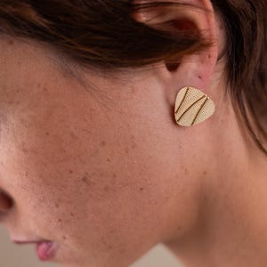 Può includere: Primo piano di un orecchino a bottone color oro di forma triangolare. L'orecchino ha una superficie strutturata con un motivo di linee sottili e parallele. L'orecchino è indossato sull'orecchio.