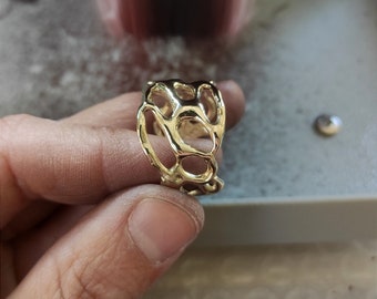 May include: A gold-colored ring with an open, organic design. The ring features a dark, rectangular stone set within the intricate, web-like structure. The ring is held in a hand, showcasing its unique, artistic style. The ring is a piece of jewelry.