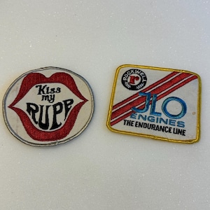 May include: Two embroidered patches. The left patch is oval-shaped with red lips and the black text "Kiss my RUPP". The right patch is rectangular with a gold border, featuring the text "ROCKWELL", "JLO ENGINES", and "THE ENDURANCE LINE".