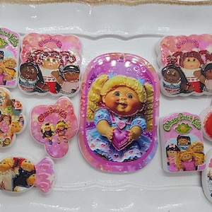 May include: A collection of nine Cabbage Patch Kids themed resin cabochons in various shapes including rectangles, ovals, hearts, and a candy shape. Each piece features colorful printed graphics of Cabbage Patch Kids characters and logos.