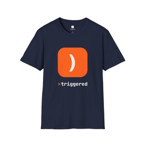 May include: A navy blue short-sleeve t-shirt featuring a graphic on the chest. The graphic consists of an orange rounded square containing a white closing parenthesis symbol, positioned above the white text that reads >triggered.
