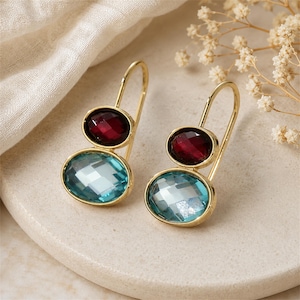 May include: A pair of gold-tone drop earrings featuring two oval-shaped faceted stones each. The top stone is a deep red, while the larger bottom stone is a light blue. The earrings are suspended from simple gold-tone wire hooks.
