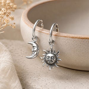 Puede incluir: Un par de pendientes de aro de plata con dijes colgantes. Un pendiente tiene un dije de media luna con cara y el otro tiene un dije de sol con cara. El metal plateado tiene un acabado oxidado para resaltar las facciones de los rostros.