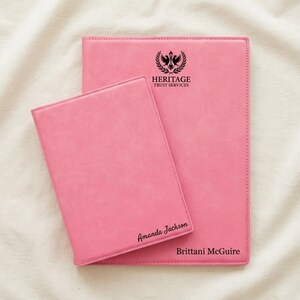 May include: Two pink faux leather folders with black text. The larger folder features a black logo with a bird and the text HERITAGE TRUST SERVICES and the name Brittani McGuire. The smaller folder features the name Amanda Jackson in black script.