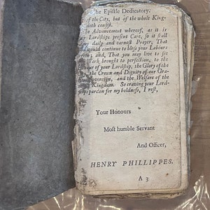 Puede incluir: Un libro antiguo con páginas envejecidas y rasgadas. El libro abierto muestra texto impreso en una fuente antigua. El texto incluye las palabras "The Epistle Dedicatory" y el nombre "HENRY PHILLIPPES."