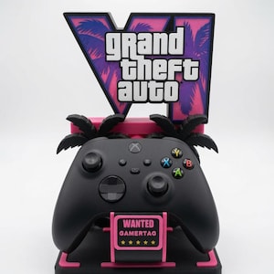 Może przedstawiać: Czarny kontroler do gier wideo umieszczony na różowo-czarnej podstawce z logo Grand Theft Auto V i sylwetkami palm. Podstawa zawiera różowy prostokątny znak z napisem WANTED GAMERTAG oraz pięcioma żółtymi gwiazdkami.