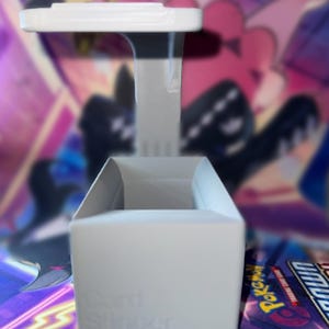 May include: A white 3D-printed card storage box with an open top and a vertical arm extending upward to a flat horizontal platform. The front of the box features the embossed text Card Stinger. The structure is rigid with a matte finish.