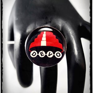 Può includere: Un anello rotondo nero con un design rosso e bianco e la scritta "DELO" in lettere bianche. L'anello è posto su uno sfondo nero.