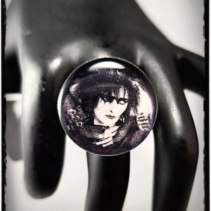 May include: A black and white photo of a person with dark hair and dark eyes. The photo is on a round, clear glass cabochon with a glittery finish. The cabochon is set in a ring.