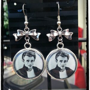 May include: Silver earrings with a bow charm and a round image of James Dean. The image is in black and white.