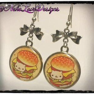 May include: A pair of silver dangle earrings featuring a cute cartoon cat peeking out from a hamburger. Each earring has a silver bow at the top.