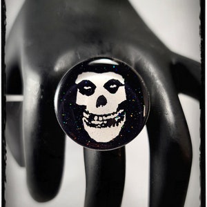Könnte beinhalten: Ein schwarzer Ring mit einem weißen Misfits-Schädel-Logo auf schwarzem Hintergrund mit Glitzer.