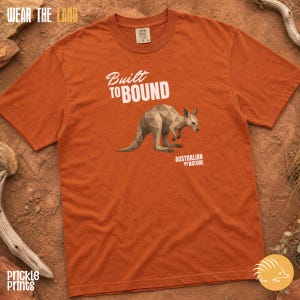 Peut inclure: Un t-shirt à manches courtes de couleur rouille avec un motif géométrique de kangourou dans des tons bruns. Le texte indique "Built TO BOUND" et "AUSTRALIAN BY NATURE" en blanc. Le t-shirt possède un col rond et une étiquette Comfort Colors.
