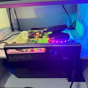Peut inclure: Une console de jeu modifiée sans son boîtier extérieur, révélant une carte mère verte et un ventilateur circulaire éclairé par des LED rouges et violettes. Le panneau avant affiche le texte MW3 et un bouton d'alimentation bleu lumineux.