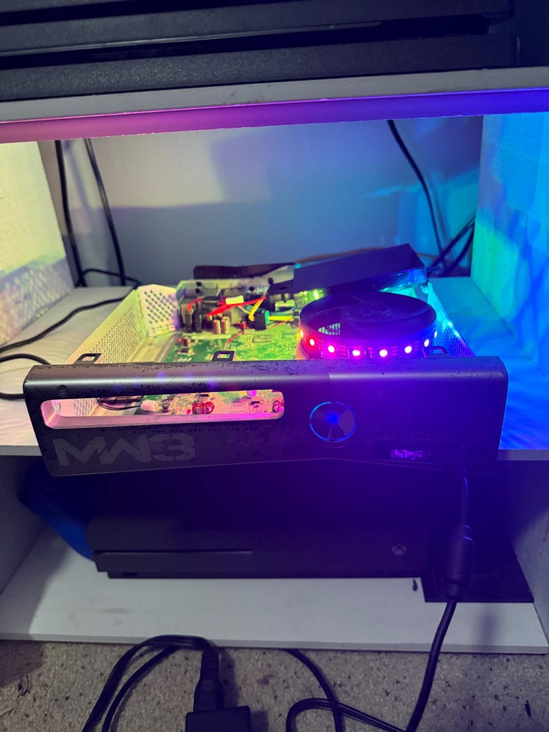 May include: A modified game console with its outer casing removed, revealing an internal green circuit board and a circular cooling fan illuminated by red and purple LED lights. The front panel features the text MW3 and a glowing blue power button.