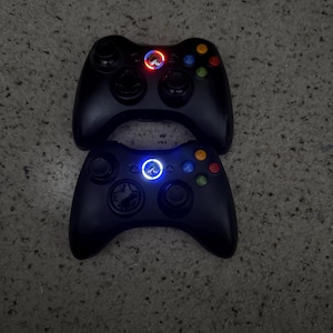 May include: Two black wireless game controllers featuring dual analog sticks, a directional pad, and four colored action buttons labeled A, B, X, and Y. The top controller displays a glowing red circular light, while the bottom controller shows a blue light.