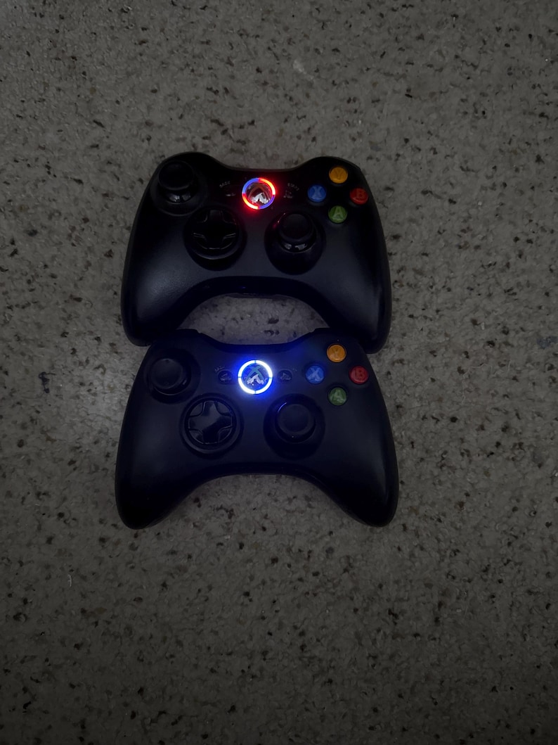May include: Two black wireless game controllers featuring dual analog sticks, a directional pad, and four colored action buttons labeled A, B, X, and Y. The top controller displays a glowing red circular light, while the bottom controller shows a blue light.