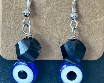 May include: A pair of drop earrings featuring silver-tone metal hooks, faceted black beads, and circular blue, white, and black evil eye charms. Each earring is finished with a small, dark, cylindrical bead at the bottom for a balanced look.