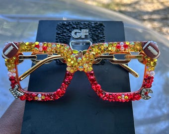 May include: Square-framed glasses embellished with red, orange, and yellow rhinestones and pearls. The frames feature miniature football charms at the top corners and silver-toned letter charms K and C at the bottom corners, with gold-toned chain-link temples.