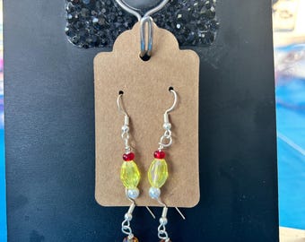 May include: A pair of dangle earrings featuring silver-toned hooks, small white and red beads, faceted yellow beads, and larger, irregular, marbled orange and yellow glass pendants. The earrings are displayed on a rectangular brown card.
