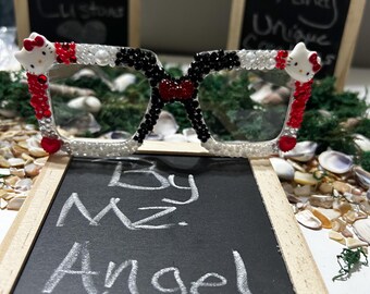 May include: Clear-framed glasses embellished with black, white, and red rhinestones, featuring two white cat character faces on the upper corners. The frames are decorated with small pearls and red heart accents along the edges of the lenses.