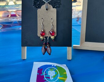 May include: Dangle earrings featuring a brown football charm with white laces, a red heart-shaped bead, and a small gold bead. The earrings are displayed on a tan card attached to a black stand with the letters GF in silver rhinestones at the top.