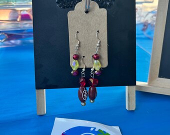 May include: A pair of dangle earrings featuring silver-toned hooks, red and yellow faceted beads, and a brown football-shaped charm with white laces. The earrings are displayed on a tan cardboard tag attached to a black stand with the letters GF.