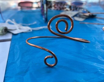 May include: A copper wire cuff bracelet featuring two open-ended spiral designs at each terminal. The metal has a polished, metallic finish with a warm reddish-brown hue. The bracelet is formed from a single continuous piece of rounded wire.