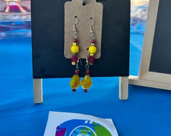 May include: A pair of dangle earrings featuring silver-tone hooks, yellow spherical beads, red faceted beads, and yellow teardrop-shaped glass beads with red swirls. The earrings are displayed on a brown card hanging from a black stand.