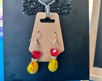 May include: A pair of dangle earrings featuring a red heart-shaped bead above a yellow teardrop-shaped bead with red swirls. Each earring includes a silver-toned metal spacer with small clear crystals and a silver-toned hook for attachment.