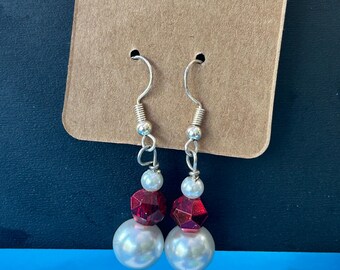 May include: A pair of drop earrings featuring silver-toned hooks, each adorned with a small white bead, a faceted translucent red bead, and a larger white spherical bead at the base, displayed on a rectangular brown cardboard jewellery card.