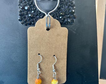 May include: A pair of drop earrings featuring silver-toned hooks and a vertical arrangement of three beads each. The beads consist of a mottled orange and yellow stone, a shiny gold-toned metallic sphere, and a smooth cream-coloured pearl-like bead.
