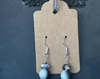 May include: A pair of dangle earrings featuring silver-toned sun charms, each suspended from a silver hook. Above the sun charm is a round, matte-finish grey bead and a smaller, faceted dark grey bead. The earrings are displayed on a brown card.
