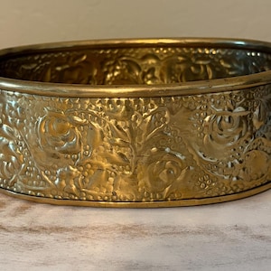 Puede incluir: Un cuenco ovalado de latón con un patrón floral en relieve de rosas y hojas en su superficie exterior. El cuenco incluye dos asas decorativas ornamentadas en los lados opuestos y tiene un acabado metálico pulido de tono dorado.