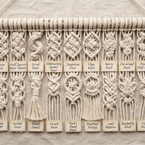 May include: A macrame wall hanging featuring nine vertical strands of off-white cord suspended from a wooden dowel. Each strand displays a unique knot pattern labeled with a small wooden tag, including Square, Spiral, Josephine, and Clove Hitch knots.