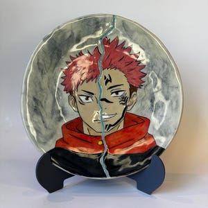 May include: A round ceramic plate featuring a hand-painted anime character with spiky red hair, a red and black hoodie, and facial markings. A vertical crack runs through the center of the plate, filled with a light blue, textured material.