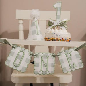 May include: A first birthday party set featuring a cone-shaped party hat with a white pom-pom, a cake topper with the number one, and a scalloped banner spelling out the word ONE. All items feature a sage green and white gingham pattern.