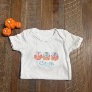 May include: A white short-sleeved bodysuit featuring an embroidered design of three orange pumpkins, each topped with a pastel-colored bow in pink, blue, and purple. Below the pumpkins, the name Klaire is embroidered in a light blue script font.