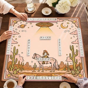 May include: A square mahjong mat featuring a desert landscape with cacti, a person riding a horse, and a UFO in the sky. The mat is light pink with brown borders and includes four racks of white mahjong tiles arranged around the perimeter.