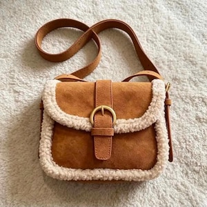 May include: A tan suede crossbody bag featuring a fold-over flap with a gold-toned metal buckle closure. The edges of the bag and the flap are trimmed with thick, cream-colored faux shearling. It includes a matching tan adjustable shoulder strap.