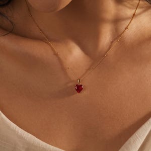 May include: A gold chain necklace featuring a small, faceted heart-shaped red gemstone pendant. The delicate chain is accented with evenly spaced gold beads, creating a refined look that rests against the collarbone with a simple, elegant design.